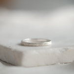 A thin silver band ring with a florentine hand engraved finish, resting on a white stone slab against a soft, blurred light gray background.