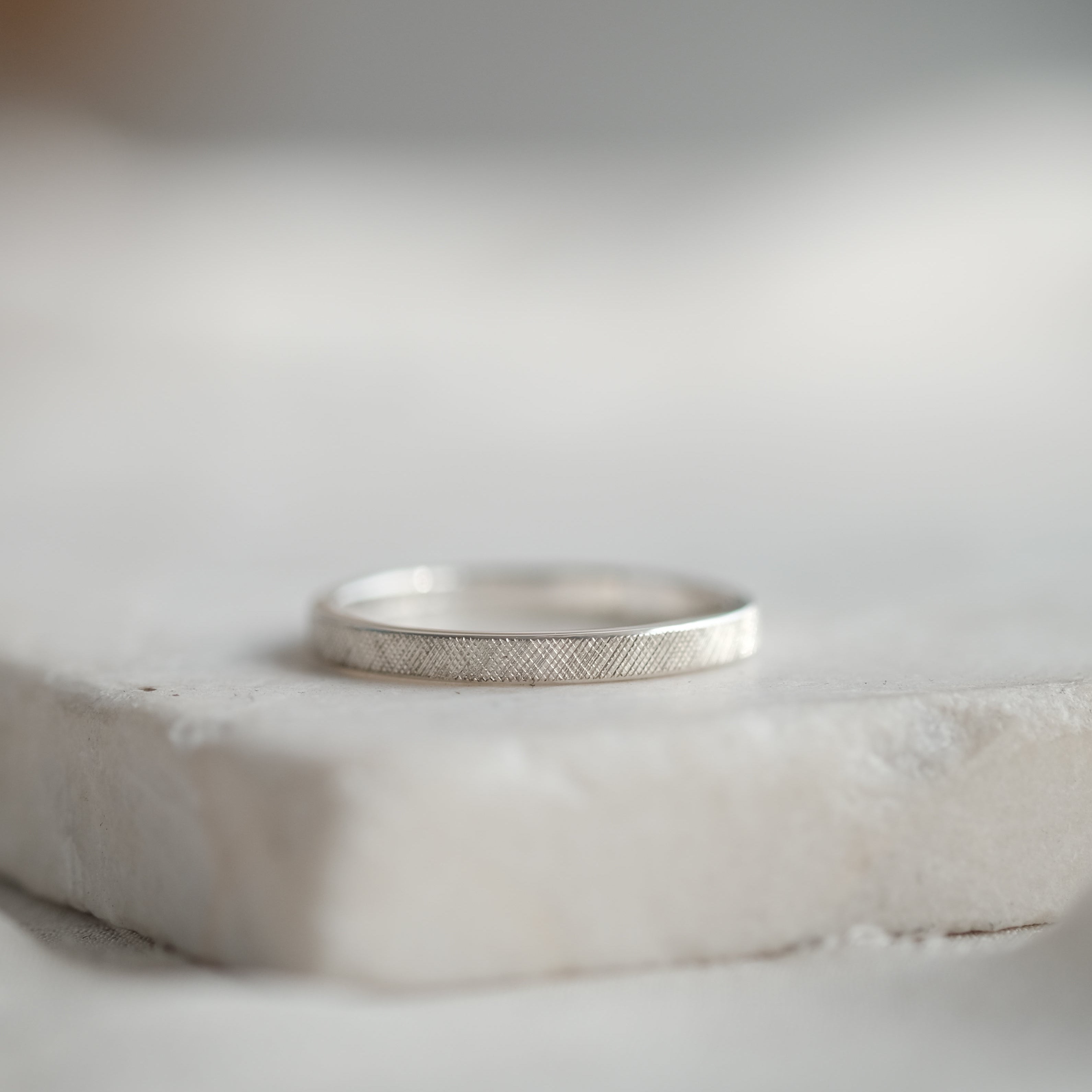 A thin silver band ring with a florentine hand engraved finish, resting on a white stone slab against a soft, blurred light gray background.